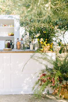 an outdoor bar with flowers and bottles on the counter, surrounded by greenery in front of it