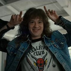 a man with long hair wearing a jean jacket and holding his hands up in the air