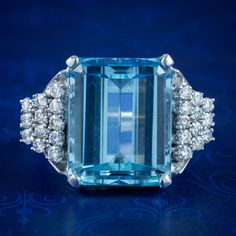 A magnificent vintage cocktail ring made in the Art Deco fashion with a colossal emerald cut aquamarine in the centre, weighing approx. 15ct with a pleasing, deep ocean blue hue.  The aqua is complemented by an audience of twenty-four sparkling brilliant cut diamonds arranged in a graduated, stepped design down each shoulder. They're 0.05ct each (approx. 1.20ct total) and have superb SI1 clarity - H colour.  The robust gallery is modelled entirely in 18ct white gold and displays the stones high Modern Blue Baguette Cut Emerald Ring, Formal Light Blue Diamond Ring, Elegant Light Blue Aquamarine Diamond Ring, Classic Light Blue Topaz Ring For Formal Occasions, Formal Aquamarine Diamond Ring With Brilliant Cut, Aquamarine Brilliant Cut Diamond Ring For Formal Occasions, Luxury Aquamarine Octagon Ring, Luxury Octagon Aquamarine Ring, Formal Light Blue Aquamarine Diamond Ring