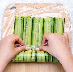 a person is holding up some cucumbers on a cutting board with plastic wrap around them