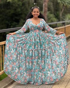 a woman standing on a bridge wearing a long dress with floral print, smiling at the camera