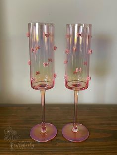 two wine glasses with pink flowers on them sitting on a wooden table next to each other