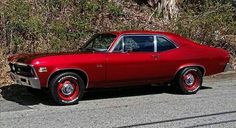 an old red car is parked on the side of the road in front of some bushes