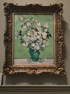 a painting with white flowers in a green vase on a wall next to a gold frame