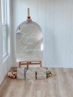 an easel with wedding signs and flowers on the floor next to it in front of a white painted wall