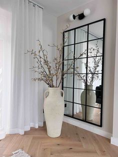 a vase with flowers in it sitting next to a large mirror on the wall near a window