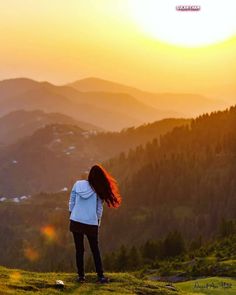 a woman standing on top of a lush green hillside next to a forest at sunset