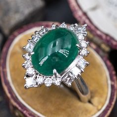 This magnificent ring is centered with one (1) oval cabochon cut natural emerald weighing 5.18 carats and set into a four-prong setting. The emerald is bordered with eight (8), prong set, square step cut diamonds and twelve (12), prong set, round brilliant cut diamonds. The ring measures 23.1mm at the top, rises 12.2mm above the finger, tapering to 3.8mm wide and 1.9mm thick at the base of the shank. This ring is currently a size 7.75. Fine Jewelry Emerald Oval Cabochon Ring, Fine Jewelry Emerald Ring With Oval Cabochon, Luxury Oval Emerald Birthstone Ring, Luxury Emerald Ring With Oval Cabochon, Wedding Emerald Ring In Oval Cabochon Shape, Elegant Emerald Ring With Cabochon Cut, Elegant Cabochon Emerald Ring, Elegant Emerald Cabochon Ring, Wedding Emerald Ring With Oval Cabochon