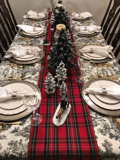 the table is set for christmas dinner with red and black plaid runneres, silver place mats, white plates, and evergreens