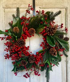 a wreath with pine cones, holly and red berries is hanging on a white door