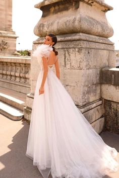 a woman in a white wedding dress is standing on the sidewalk near a stone wall