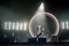 a man on stage playing an electric guitar with other people in the background and lights behind him