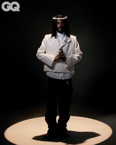 a man wearing a white suit and crown standing in front of a black background with the word gq on it