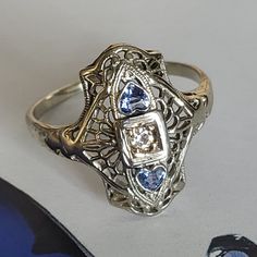 "Art Deco Navette Diamond Sapphire Filigree Ring. This piece is tested 18K white gold cast fashion lady's ring sizeable 7 1/2 measuring 18.35.. x 13.66mm with bright polish finish weighting 2.79 grams. The featured ring is Navette style and delicate filigree design rectangular round corners curved top with pierced out filigree and milgrain details centered by a square double bezel box holding a prong set round Old Mine cut Diamond measuring 2.55mm approx. 0.10ct guarded by each side with a pear Art Deco Sapphire Ring, Art Deco Filigree, Horseshoe Ring, Estate Ring, 10k Gold Ring, Old Mine Cut Diamond, Estate Rings, Pacific Palisades, Gold Art Deco