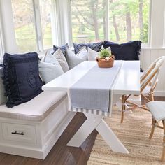 a white table and bench with blue pillows on it in front of a bay window