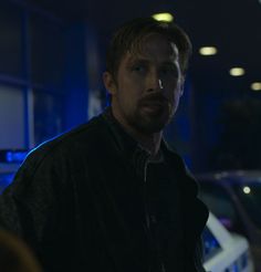 a man in a black jacket standing next to a police car and looking at the camera