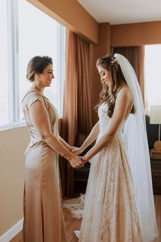 two women standing next to each other in front of a window wearing wedding gowns