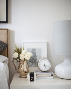 a nightstand with a lamp, clock and flowers on it next to a framed photograph