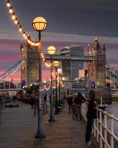 people are walking on the sidewalk by the water at dusk with lights strung across the walkway