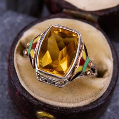 This fun ring features a checkerboard rectangular cut citrine flanked by yellow, black, green, and red enamel accented shoulders. The ring is crafted in 10k white gold and is currently a size 5.5. Due to the enamel details, resizing options are limited. Luxury Vintage Gold Enamel Ring, Art Deco Enamel Ring As A Gift, Art Deco Enamel Ring As Gift, Collectible Hallmarked Enamel Ring In Fine Jewelry Style, Art Deco Enamel Ring Jewelry, Art Deco Enamel Jewelry Ring, Retro Collectible Enamel Jewelry, Art Deco Multicolor Jewelry For Gift, Multicolor Art Deco Jewelry For Gift