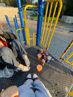 two people sitting on a playground with drinks and snacks in front of them, one person has their feet up on the ground