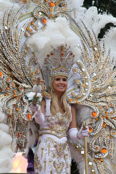 a woman in a white and gold costume with feathers on her head, holding a stick