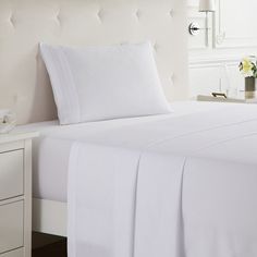 a bed with white sheets and pillows in a bedroom next to a night stand that has flowers on it