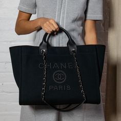 This Chanel Large Deauville Tote is the perfect elevated casual design from Chanel's spring/summer 2020 bag collection. A thick black canvas exterior, referred to as 'mixed fibers' by Chanel, makes up the exterior of this pristine bag, accented by black leather trim and silver-tone hardware. Front and center, the Chanel lettering and logo present in a black faux pearl design, drawing in attention with a hint of shine. Carry this Chanel tote with the dual rolled leather top handles, or wear it on your shoulder with the interwoven leather & chain shoulder straps with a leather comfort guard. The top of this 'it bag' secures via one magnetic snap closure, leading into the black canvas interior. A spacious design, inside you will find one zip pocket along the back wall, two slip pockets, two o Luxury Black Rectangular Canvas Bag, Luxury Canvas Bag With Top Handle, Luxury Large Capacity Canvas Bag, Luxury Large Capacity Canvas Bag For Shopping, Luxury Large Capacity Canvas Shopping Bag, Luxury Black Canvas Bag For Everyday, Black Shopping Bag With Canvas Lining, Designer Black Canvas Shoulder Bag, Luxury Everyday Canvas Bag