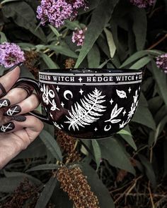 a hand holding a black and white coffee mug in front of purple flowers with writing on it