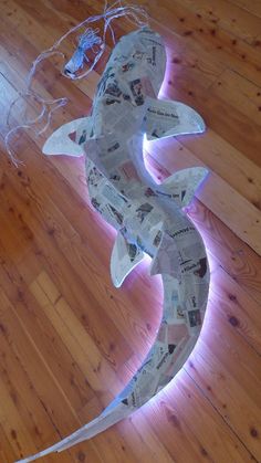 a fish made out of newspaper sitting on top of a wooden floor next to a light