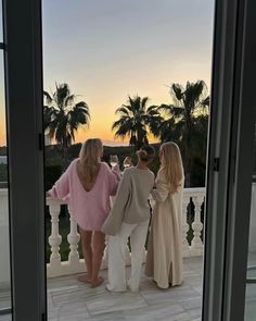 three women standing on a balcony looking out at the sunset and palm trees in the distance