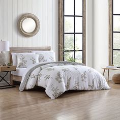 a bed with white comforter and pillows in a room next to two large windows