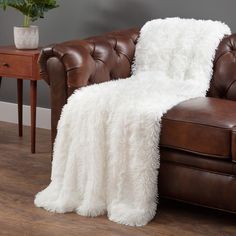 a brown leather chair with a white fluffy blanket on it's back next to a wooden table