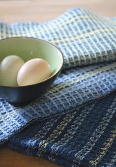 three eggs in a bowl on top of a table cloth next to two blue yarn swatches