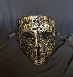 This men's Venetian masquerade mask is made of a lightweight metal and is hand painted in gold and black. The abstract paint job gives the mask a unique look. Stand out at your next costume or masquerade party with this awesome mask! Mask has some flexibility and come with black silk ribbon ties attached. Mask size 6"W x 7"H. Mask color gold/black Gold Fantasy Festival Masks And Prosthetics, Gold Masks And Prosthetics For Carnival Festival, Gold Masquerade Mask For Mardi Gras Festival, Gold Steampunk Costume Accessories For Parties, Gold Steampunk Costume Accessories For Costume Party, Gothic Gold Costume Accessories For Masquerade, Gothic Gold Costume Accessories For Costume Party, Gold Gothic Costume Accessories For Costume Party, Gold Steampunk Costume Accessories For Festival