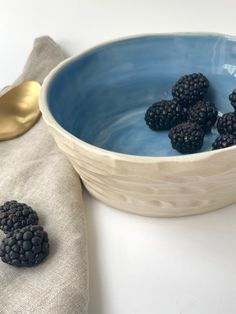 some blackberries are in a blue bowl next to a gold spoon and linen napkin