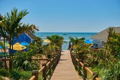 a wooden walkway leading to the beach with umbrellas and palm trees on either side