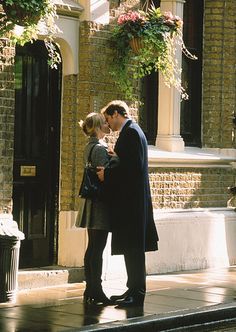 a man and woman standing on the sidewalk in front of a building with potted plants