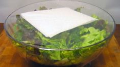 a glass bowl filled with lettuce sitting on top of a wooden table next to a white napkin