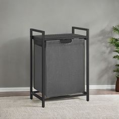 a grey trash can with a black frame on the floor next to a potted plant