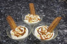 three desserts in small glass bowls with cinnamon sticks sticking out of the top and caramel sauce drizzled on them