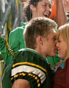 a man and woman kissing in the rain with other people behind them smiling at each other