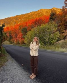 a woman standing on the side of a road in front of trees with fall colors