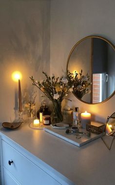 a white dresser topped with lots of candles and vases filled with flowers next to a round mirror