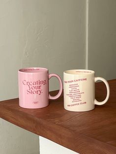 two coffee mugs sitting on top of a wooden table next to each other, one pink and one white