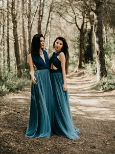 two women in blue dresses standing on a dirt road with trees and bushes behind them