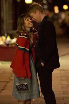 a man and woman standing next to each other on a sidewalk at night with lights in the background