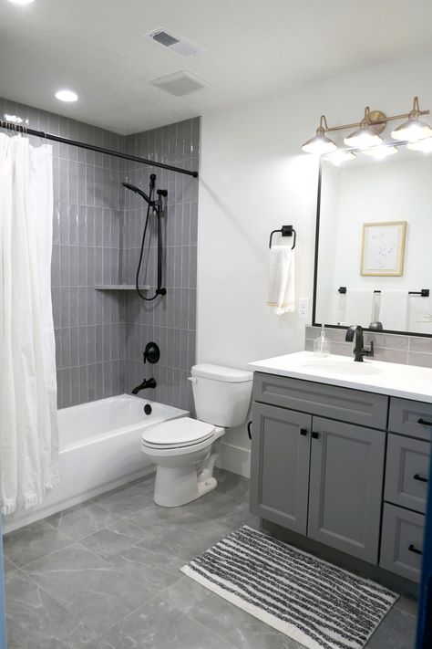 A bathroom with a tub/shower combo that has tub to ceiling gray glass subway tile installed vertically. The toilet is between the tub and single-sink vanity, which has darker gray cabinets and white Quartz countertops. The floor has large rectangular tiles that are gray with white veins. White And Dark Grey Bathroom, White Bathroom With Grey Floor, Grey Bathroom Styling, Modern Bathroom Grey And White, Small Bathroom With Grey Vanity, Bathroom Gray And Black, Gray Toilet Bathroom, Grey Shower Tile Bathroom, Sink Toilet Shower Bathroom Layout