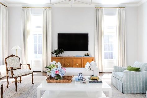 White curtains hang from brass curtain rods in front of windows flanking wooden TV console located beneath a wall mount TV. Tv Between Windows, Windows Design Ideas, Brown Wooden Bed, White Settee, Windows Design, Blue And White Pillows, Tv Wall Decor, Cottage Living Rooms, Transitional Living