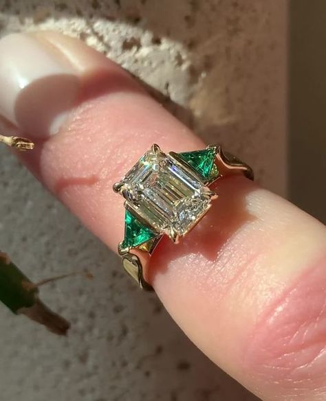 Frank Darling Custom Rings on Instagram: "Send this to someone who loves gemstone accents 💎💚⁠ ⁠ This emerald three stone engagement ring is all custom from head to toe. Let's break it down:⁠ ⁠ • Lab grown green emerald trillion side stones⁠ • 2.1ct, D Color, VVS2 Center Emerald⁠ Diamond • 3.5mm Square band that tapers into cathedral shoulders⁠ • Our signature Breezy setting, to top it off ✨⁠ • All set in a shiny 18k Yellow Gold⁠ ⁠ Mark your calendars, darlings! The last day for Christmas proposal orders is December 1st 💍❄️⁠ ⁠ #emeralddiamondengagementring #emeraldengagementring #threestonering #threestoneengagementring #emeraldcutdiamond #engagementringinspiration #engagmentringideas #gemstonering #customengagementring⁠ ⁠ 𝐖𝐚𝐧𝐭 𝐭𝐨 𝐠𝐞𝐭 𝐬𝐭𝐚𝐫𝐭𝐞𝐝 𝐝𝐞𝐬𝐢𝐠𝐧𝐢𝐧𝐠 𝐲𝐨𝐮𝐫 ? Engagement Ring With Emerald Side Stones, Emerald Ring With Gold Band, Engagement Rings Diamond And Emerald, Emerald Ring With Diamond Band, Emerald Side Stone Engagement Ring, Emerald Accent Engagement Ring, Yolanda Diaz Wedding Ring, Emerald Gemstone Ring, Vintage Yellow Gold Emerald Ring